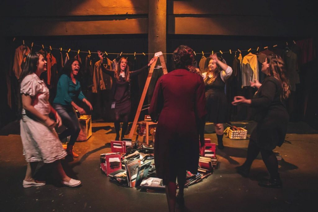 Seis integrantes do Grupo Alumiah Teatro, todas mulheres, encenando uma peça teatral numa sala com pouca iluminação, com um varal cheio de roupas ao fundo e ao centro da sala uma escada aberta rodeada de livros.