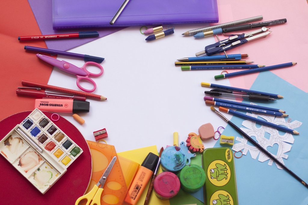 Em uma mesa, material escolar disperso: folhas coloridas, e por cima, aquarela, lápis, canetas, tesouras...