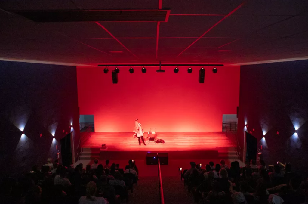 Foto do fundo do cine teatro, mostrando parte da plateia na parte inferior, e todo o restante da cena, em vermelho, o palco e as paredes, durante uma apresentação de teatro.