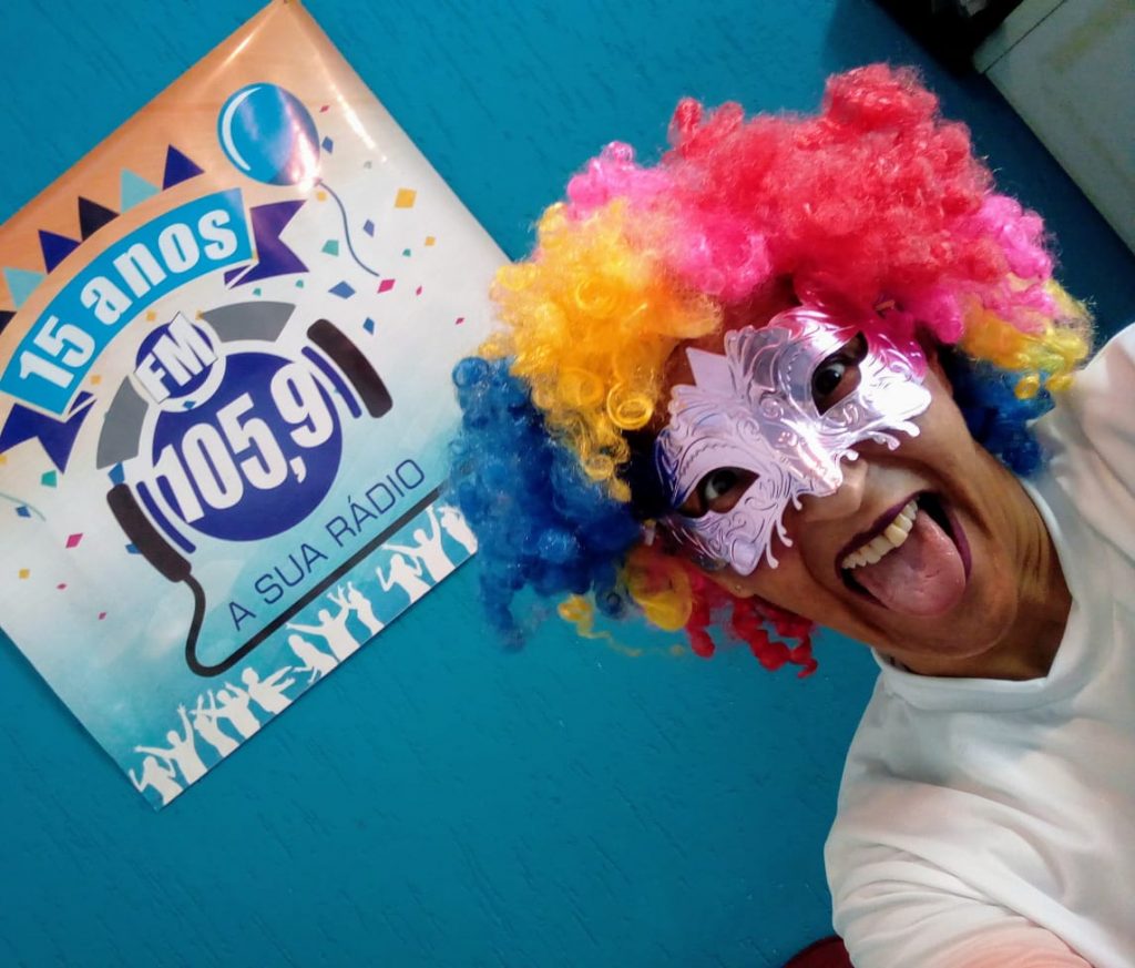 Glaucia com uma máscara e peruca colorida, de carnaval, com a língua de fora, na frente de um dos cartazes da rádio.