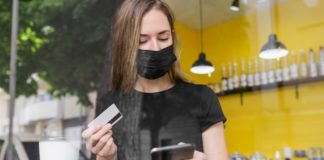 Comprar em Bauru. Na foto, uma mulher com uma roupa e uma máscara preta segura um cartão e um celular.