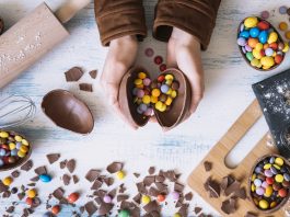 Nutricionistas de Bauru dão dicas para manter uma alimentação saudável na Páscoa Foto de um fundo branco com chocolate por cima e um par de mãos segurando um ovo de páscoa. Matéria Alimentação saudável na Páscoa: nutricionistas de Bauru dão dicas e fazem recomendações!